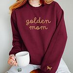 Woman holding a mug in a Maroon crewneck sweatshirt embroidered with golden mom in gold script and a golden retriever silhouette on the cuff