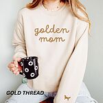 Woman holding a mug in a Sand crewneck sweatshirt embroidered with golden mom in gold script and a golden retriever silhouette on the cuff