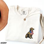 Ivory T-shirt folded beside a hat with a sitting golden retriever wearing an American flag embroidered on the left chest