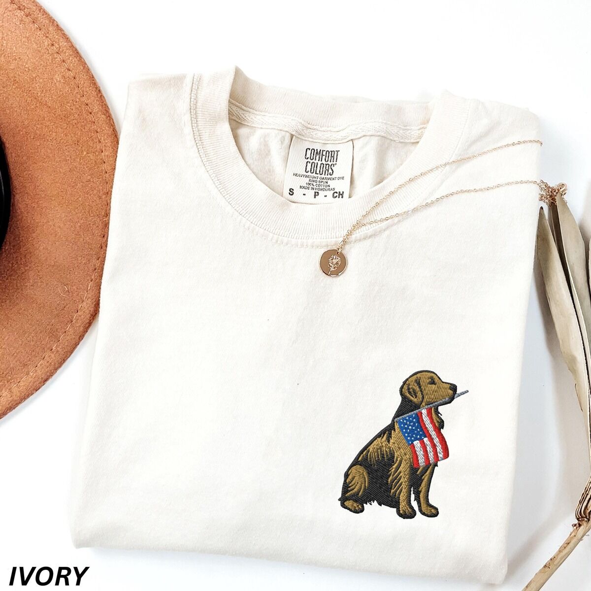 Golden Retriever American Flag Shirt – Ivory Ivory T-shirt folded beside a hat with a sitting golden retriever wearing an American flag embroidered on the left chest