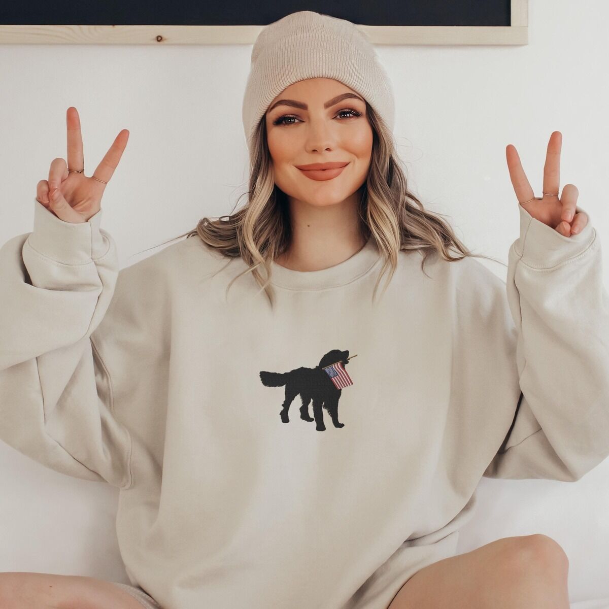 Golden Retriever American Flag Sweatshirt - worn in sand Woman in a beanie making peace signs in a sand crewneck sweatshirt with a golden retriever carrying a flag