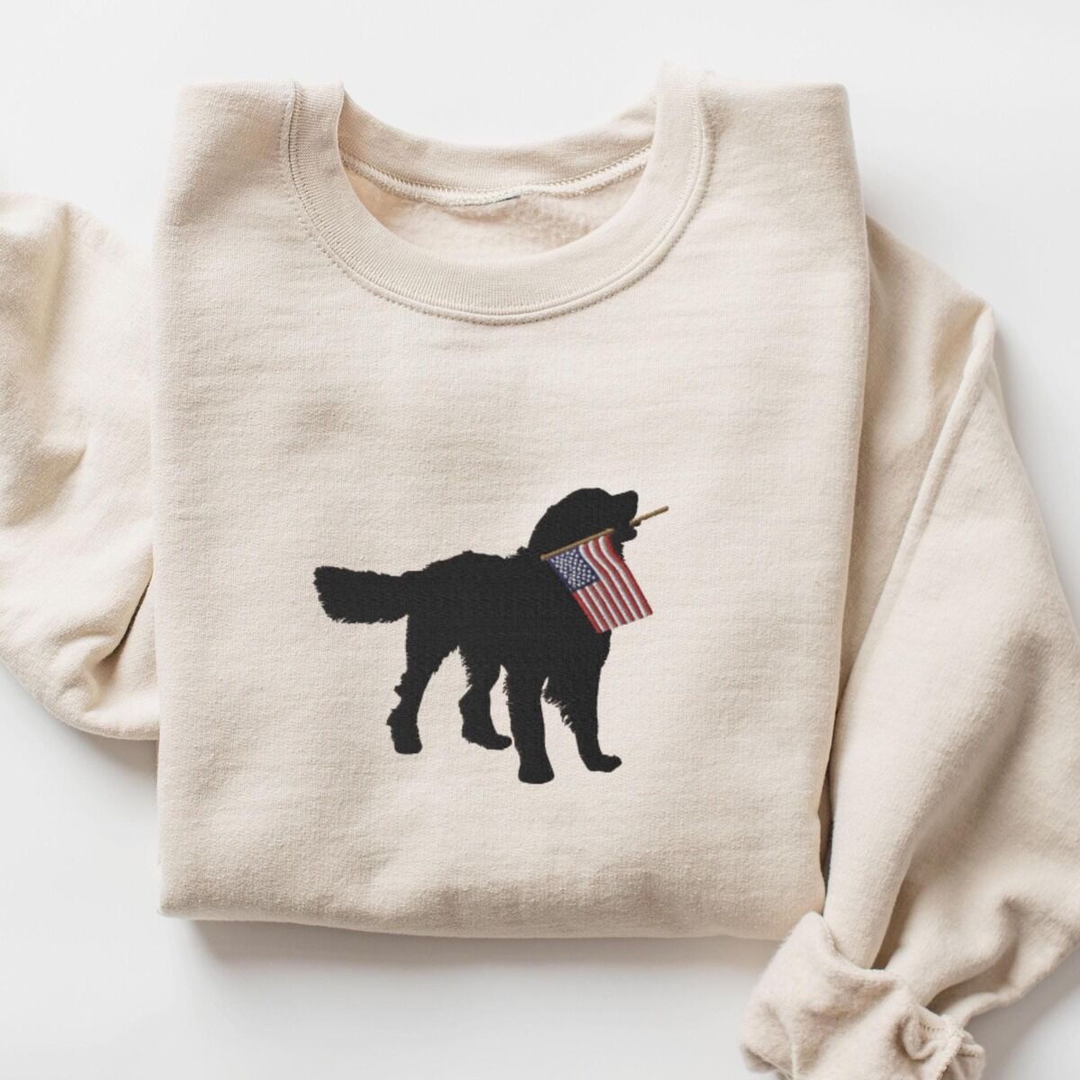 Golden Retriever American Flag Sweatshirt – Sand Sand crewneck sweatshirt laid flat with a rolled sleeve, embroidered with a golden retriever silhouette carrying a USA flag