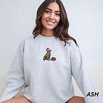 Woman sitting in an Ash crewneck sweatshirt embroidered with a golden retriever in a red Santa hat and a wrapped Christmas present