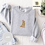 Ash crewneck sweatshirt folded with denim shorts, white sneakers and a potted plant, embroidered with a golden retriever in a Santa hat holding Christmas lights in the center of the chest