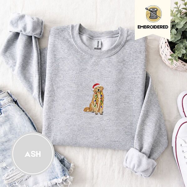 Ash crewneck sweatshirt folded with denim shorts, white sneakers and a potted plant, embroidered with a golden retriever in a Santa hat holding Christmas lights in the center of the chest