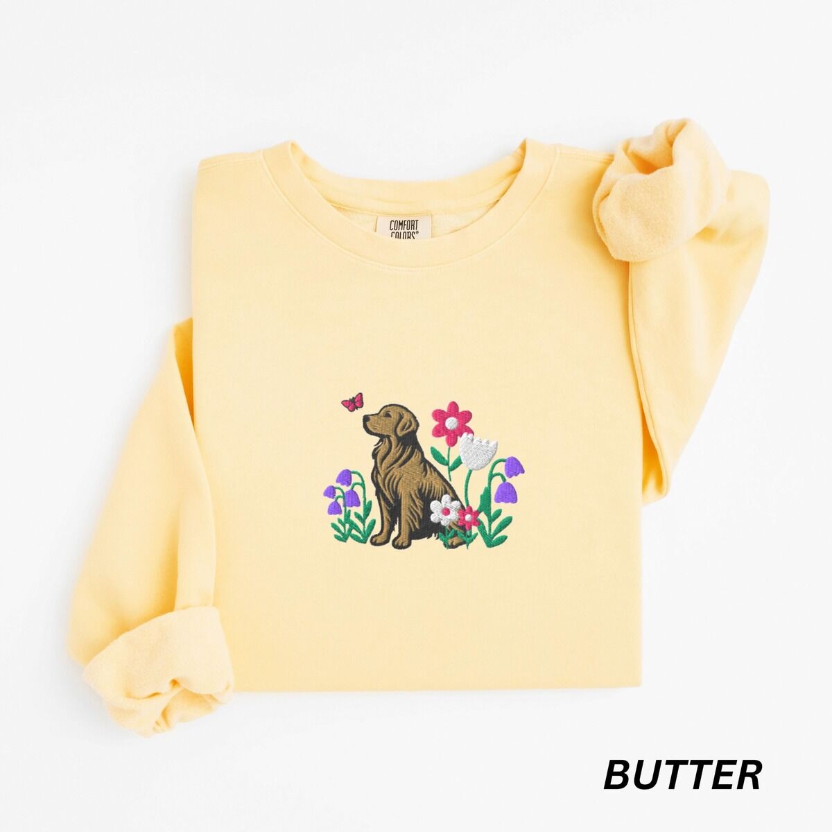Golden Retriever Flower Sweatshirt – Butter Butter crewneck sweatshirt laid flat with a rolled cuff, embroidered with a sitting golden retriever among pink daisies, a white tulip and purple bellflowers