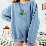Woman in an oversized Blue Jean crewneck sweatshirt embroidered with a sitting golden retriever among spring flowers