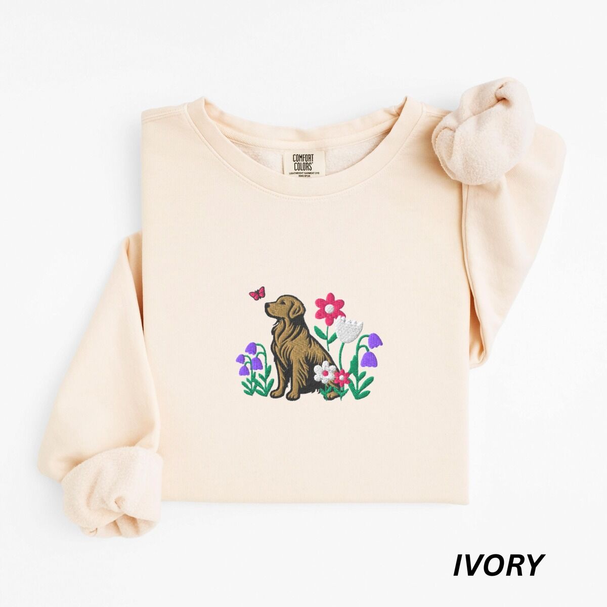 Golden Retriever Flower Sweatshirt – Ivory Ivory crewneck sweatshirt laid flat, embroidered with a sitting golden retriever among spring flowers and a pink butterfly
