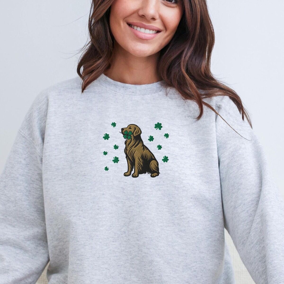 Golden Retriever Four Leaf Clover Sweatshirt – Ash Smiling woman in a Ash crewneck sweatshirt embroidered with a sitting golden retriever holding a four leaf clover in its mouth, ringed by green shamrocks