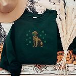 Forest Green crewneck sweatshirt laid flat with a straw hat and dried pampas, embroidered with a sitting golden retriever holding a four leaf clover in its mouth, ringed by green shamrocks
