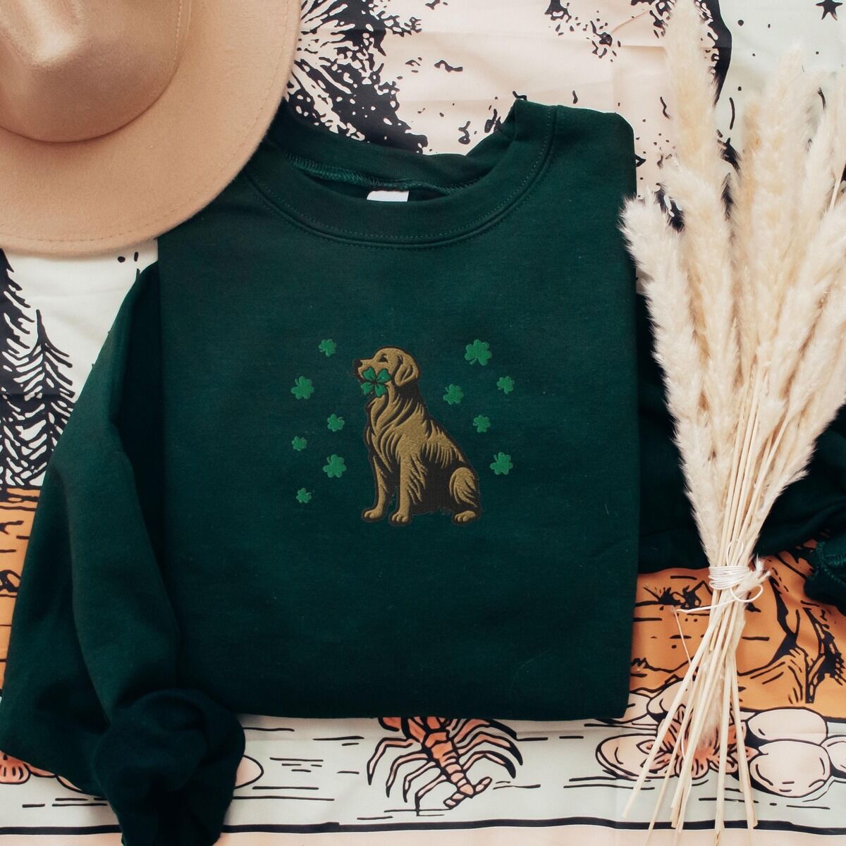 Golden Retriever Four Leaf Clover Sweatshirt – Forest Green Forest Green crewneck sweatshirt laid flat with a straw hat and dried pampas, embroidered with a sitting golden retriever holding a four leaf clover in its mouth, ringed by green shamrocks