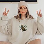 Woman in a beanie making peace signs in a Sand crewneck sweatshirt embroidered with a sitting golden retriever holding a four leaf clover in its mouth, ringed by green shamrocks