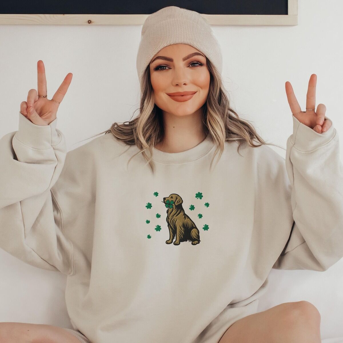 Golden Retriever Four Leaf Clover Sweatshirt - worn Woman in a beanie making peace signs in a Sand crewneck sweatshirt embroidered with a sitting golden retriever holding a four leaf clover in its mouth, ringed by green shamrocks
