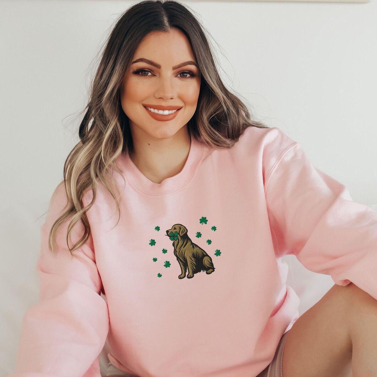 Golden Retriever Four Leaf Clover Sweatshirt - Light Pink Smiling woman in a Light Pink crewneck sweatshirt embroidered with a sitting golden retriever holding a four leaf clover in its mouth, ringed by green shamrocks