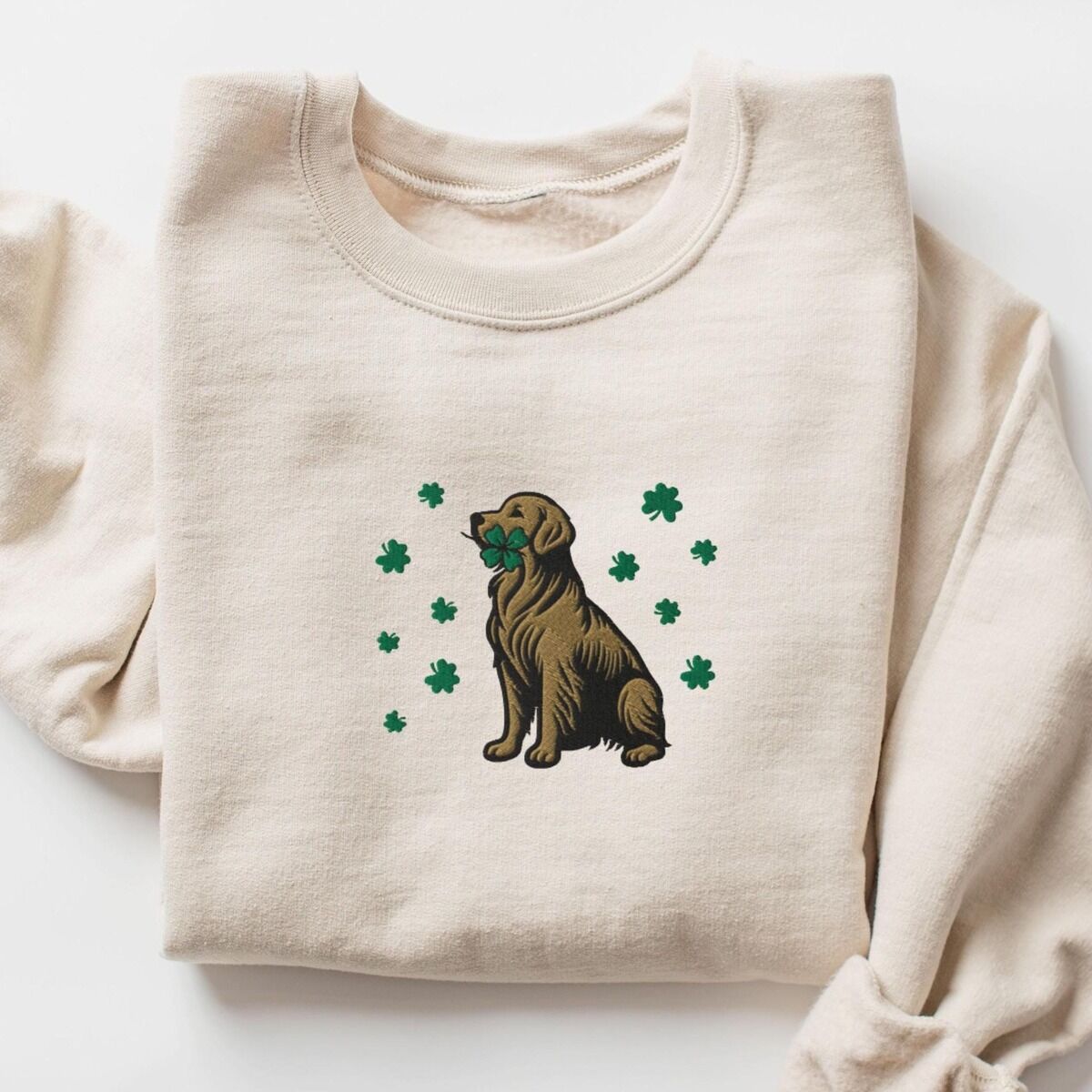 Golden Retriever Four Leaf Clover Sweatshirt – Sand Sand crewneck sweatshirt laid flat, embroidered with a sitting golden retriever holding a four leaf clover in its mouth, ringed by green shamrocks