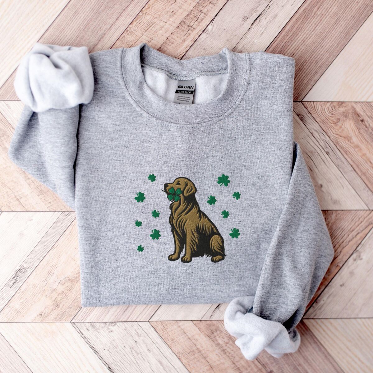 Golden Retriever Four Leaf Clover Sweatshirt - Sport Grey Sport Grey crewneck sweatshirt laid flat on a wood floor, embroidered with a sitting golden retriever holding a four leaf clover in its mouth, ringed by green shamrocks