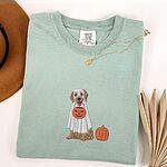 Bay T-shirt folded flat with a necklace and a brown hat, embroidered with a golden retriever in a white ghost sheet carrying a jack-o'-lantern trick-or-treat bucket beside a small pumpkin