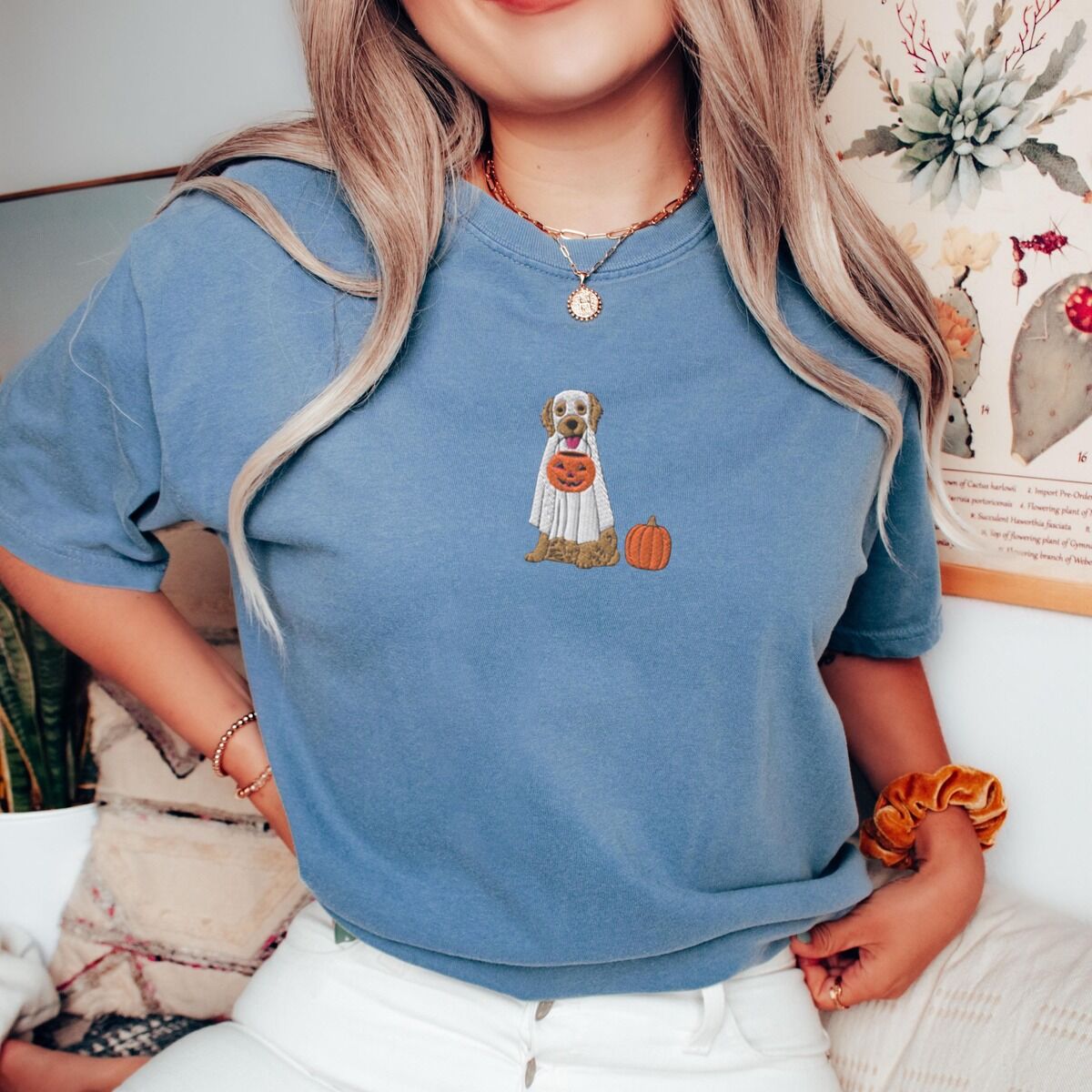 Golden Retriever Ghost Shirt – Blue Jean Woman sitting in a Blue Jean T-shirt embroidered with a golden retriever in a white ghost sheet carrying a jack-o'-lantern trick-or-treat bucket beside a small pumpkin
