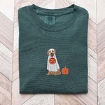 Blue Spruce T-shirt folded flat on a light wood floor, embroidered with a golden retriever in a white ghost sheet carrying a jack-o'-lantern trick-or-treat bucket beside a small pumpkin