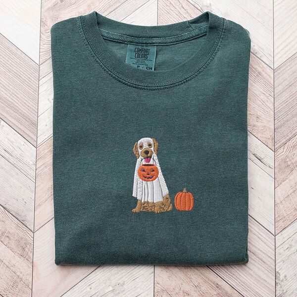Blue Spruce T-shirt folded flat on a light wood floor, embroidered with a golden retriever in a white ghost sheet carrying a jack-o'-lantern trick-or-treat bucket beside a small pumpkin