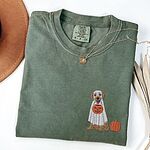 Moss T-shirt folded flat with a necklace and a brown hat, embroidered with a golden retriever in a white ghost sheet carrying a jack-o'-lantern bucket beside a small pumpkin on the left chest