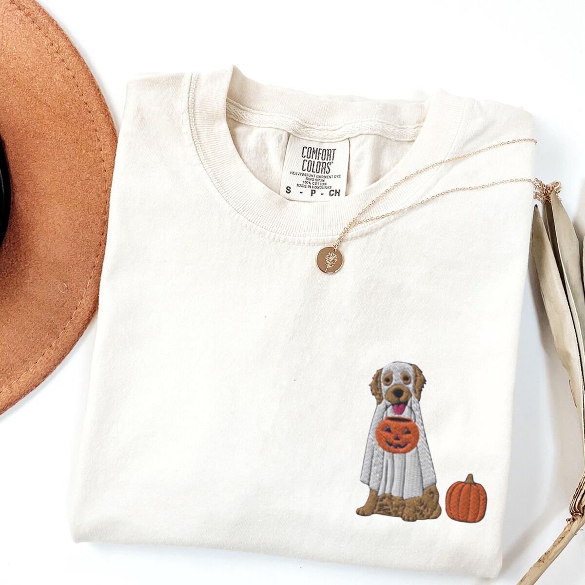 Golden Retriever Ghost Shirt - left chest on Ivory Ivory T-shirt folded flat with a necklace and dried palm leaves, embroidered with a golden retriever in a white ghost sheet carrying a jack-o'-lantern bucket beside a small pumpkin on the left chest