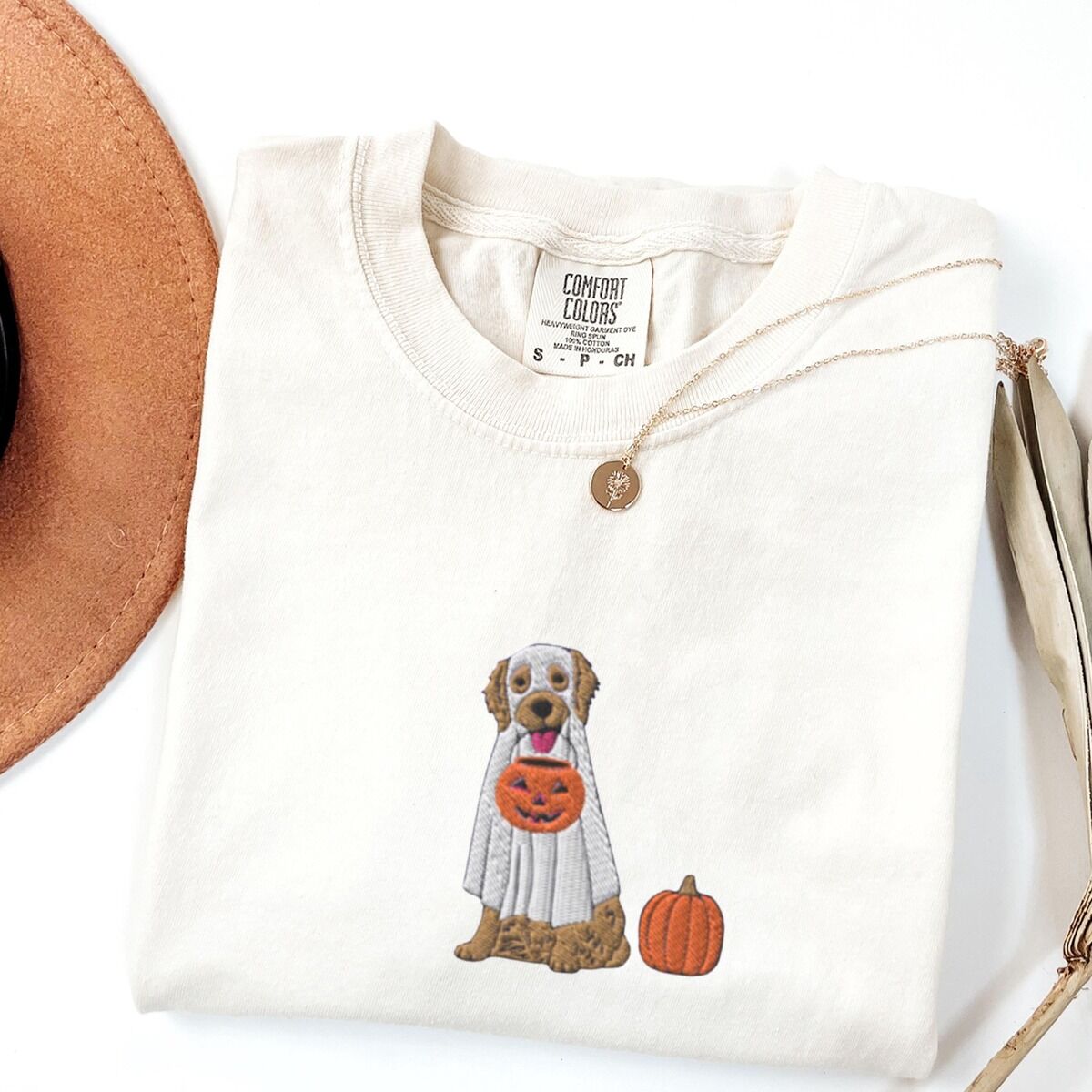 Golden Retriever Ghost Shirt – Ivory Ivory T-shirt folded flat with a necklace and dried palm leaves, embroidered with a golden retriever in a white ghost sheet carrying a jack-o'-lantern trick-or-treat bucket beside a small pumpkin