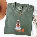 Moss T-shirt folded flat with a necklace, a brown hat and dried palm leaves, embroidered with a golden retriever in a white ghost sheet carrying a jack-o'-lantern trick-or-treat bucket beside a small pumpkin