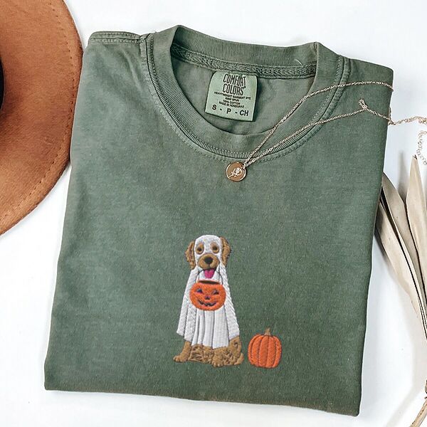 Moss T-shirt folded flat with a necklace, a brown hat and dried palm leaves, embroidered with a golden retriever in a white ghost sheet carrying a jack-o'-lantern trick-or-treat bucket beside a small pumpkin