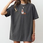 Woman in an oversized Pepper T-shirt embroidered with a golden retriever in a white ghost sheet carrying a jack-o'-lantern trick-or-treat bucket beside a small pumpkin