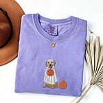Violet T-shirt folded flat with a necklace and a brown hat, embroidered with a golden retriever in a white ghost sheet carrying a jack-o'-lantern trick-or-treat bucket beside a small pumpkin