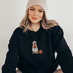 Woman in a beanie wearing a Black crewneck sweatshirt embroidered with a golden retriever in a white ghost sheet carrying a jack-o'-lantern trick-or-treat bucket beside a small pumpkin