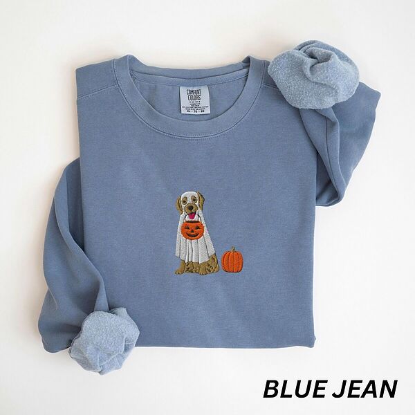 Blue Jean crewneck sweatshirt folded flat with a rolled cuff, embroidered with a golden retriever in a white ghost sheet carrying a jack-o'-lantern trick-or-treat bucket beside a small pumpkin