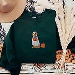 Forest Green crewneck sweatshirt laid flat with a straw hat and dried pampas, embroidered with a golden retriever in a white ghost sheet carrying a jack-o'-lantern trick-or-treat bucket beside a small pumpkin