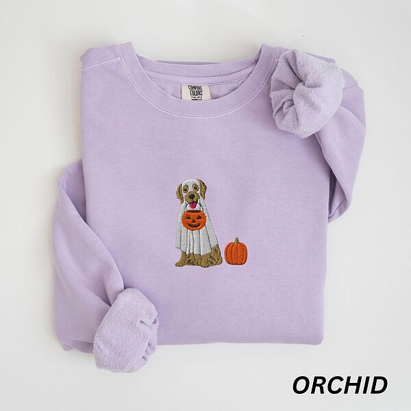 Orchid crewneck sweatshirt folded flat with a rolled cuff, embroidered with a golden retriever in a white ghost sheet carrying a jack-o'-lantern trick-or-treat bucket beside a small pumpkin
