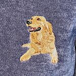 Close-up of a real stitched golden retriever lying down with its head up and tongue out, in layers of gold, honey and cream thread on a blue sweatshirt
