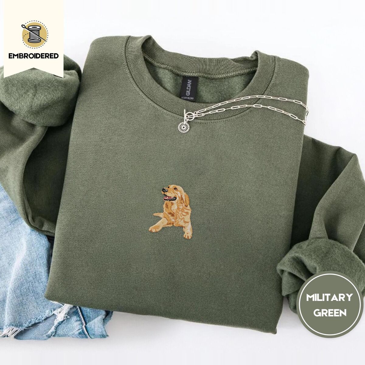 Golden Retriever Mom Sweatshirt – Military Green Military Green crewneck sweatshirt folded with light jeans and a silver chain necklace, embroidered with a happy golden retriever lying down with its head up and tongue out