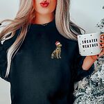 Woman holding a mug in a Black crewneck sweatshirt embroidered with a small golden retriever in a red Santa hat on the left chest