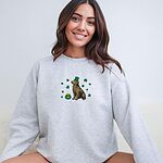 Smiling woman in a Ash crewneck sweatshirt embroidered with a sitting golden retriever in a green leprechaun hat with a pot of gold and green shamrocks