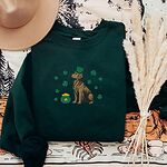 Forest Green crewneck sweatshirt laid flat with a straw hat and dried pampas, embroidered with a sitting golden retriever in a green leprechaun hat with a pot of gold and green shamrocks