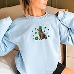 Smiling woman in a Light Blue crewneck sweatshirt embroidered with a sitting golden retriever in a green leprechaun hat with a pot of gold and green shamrocks