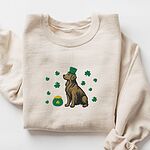 Sand crewneck sweatshirt laid flat, embroidered with a sitting golden retriever in a green leprechaun hat with a pot of gold and green shamrocks