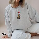 Smiling woman in a Ash crewneck sweatshirt embroidered with a sitting golden retriever holding the string of a red heart balloon in its mouth