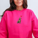 Smiling woman in a Heliconia crewneck sweatshirt embroidered with a sitting golden retriever holding the string of a red heart balloon in its mouth
