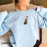 Smiling woman in a Light Blue crewneck sweatshirt embroidered with a sitting golden retriever holding the string of a red heart balloon in its mouth