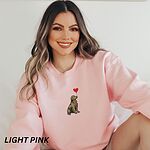 Smiling woman in a Light Pink crewneck sweatshirt embroidered with a sitting golden retriever holding the string of a red heart balloon in its mouth