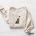 Sand crewneck sweatshirt laid flat, embroidered with a sitting golden retriever holding the string of a red heart balloon in its mouth