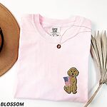 Blossom pink T-shirt folded beside a hat, a sitting goldendoodle with a USA flag stitched on the left chest