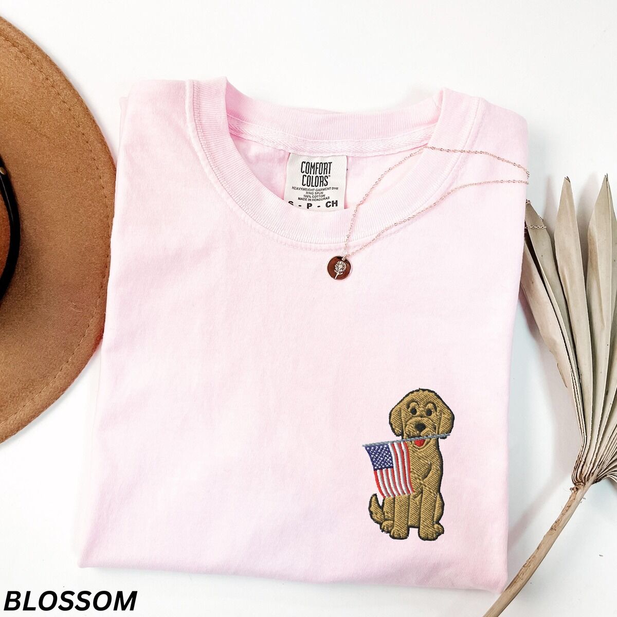 Goldendoodle American Flag Shirt – Blossom Blossom pink T-shirt folded beside a hat, a sitting goldendoodle with a USA flag stitched on the left chest