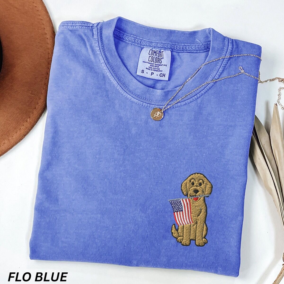 Goldendoodle American Flag Shirt – Flo Blue Flo Blue T-shirt folded beside a hat with a curly goldendoodle holding an American flag embroidered on the left chest